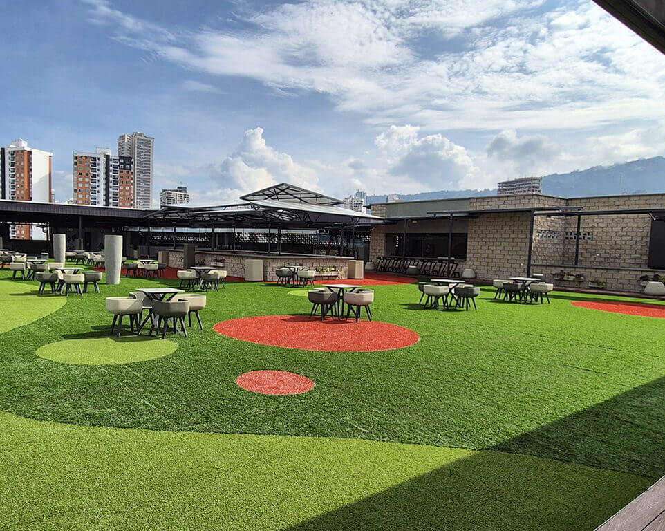 Terraza al aire libre con césped sintético, mesas y sillas distribuidas para actividades sociales o eventos, con edificios urbanos al fondo bajo un cielo despejado.