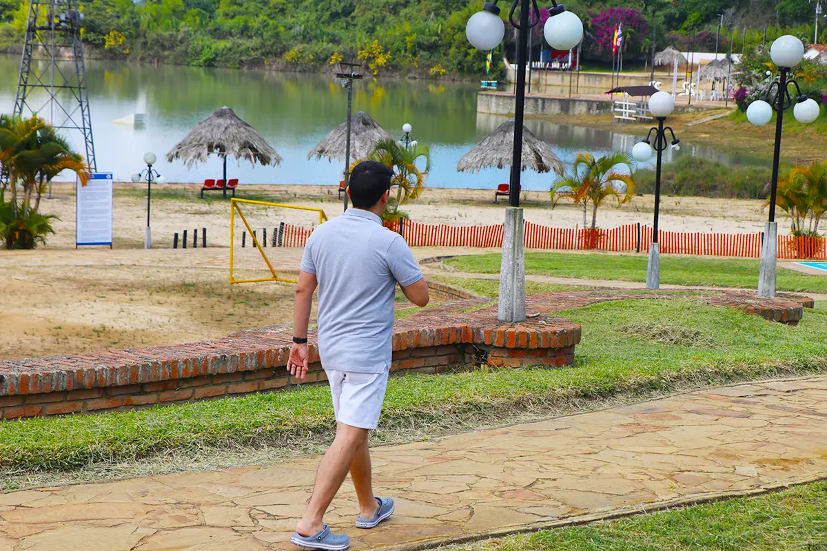 Hombre camina por sendero con lago, palmeras y kioscos de paja en el parque Acuarela Cajasan