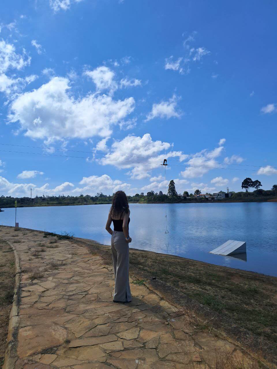 Mujer de espaldas observando el lago de Acuarela Cajasan, con cielo azul, sendero de piedra y cable para deportes acuáticos